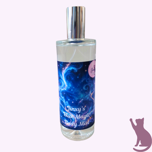 A clear glass bottle of body mist with a silver cap and a blue galaxy-themed label reading 'Jinxy's Blue Magic Body Mist', shown on a pale pink background with a purple cat silhouette in the lower right corner.