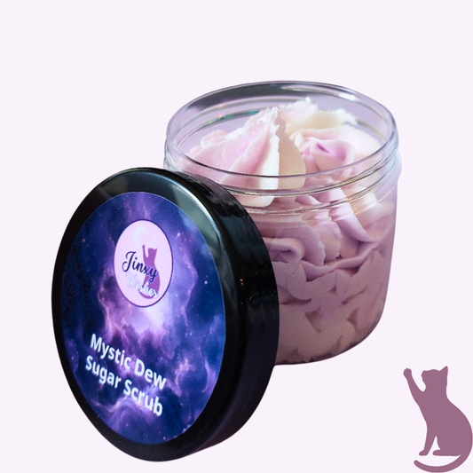 A jar of Jinxes Mystic Dew Sugar Scrub with its black lid featuring a purple galaxy design leaning against the jar, set against a pale pink background.